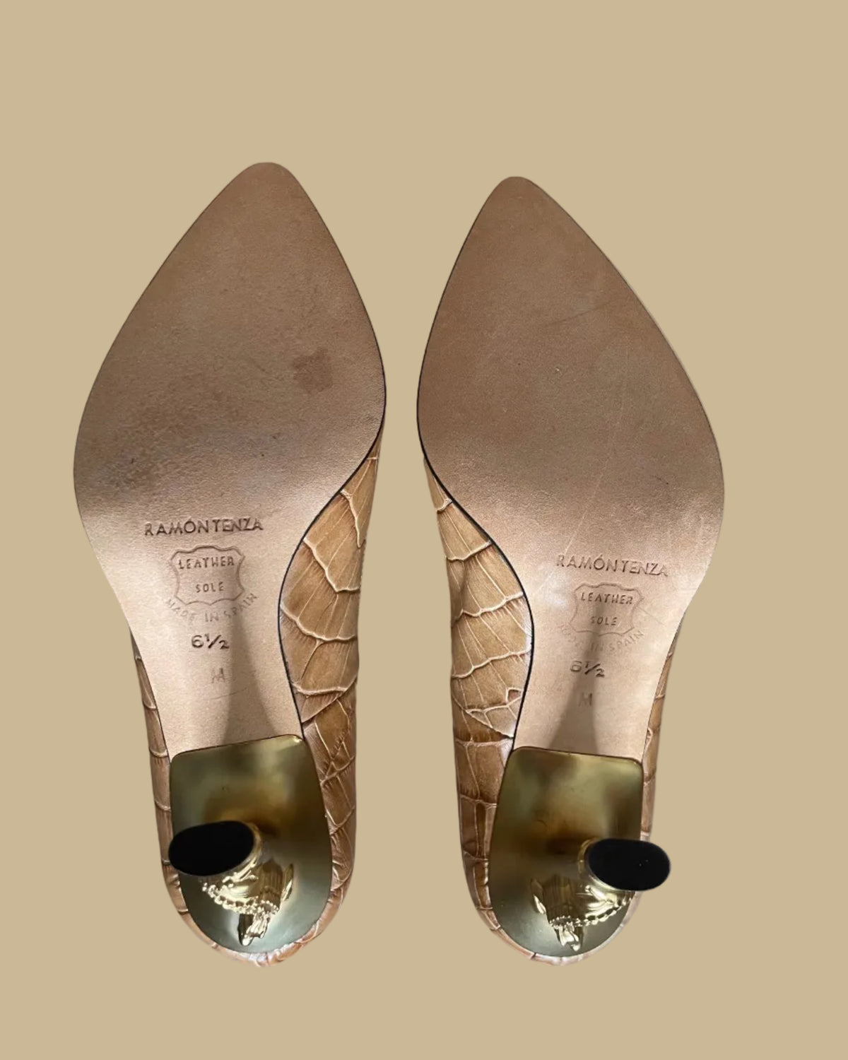 Top-down view of a pair of pointed-toe high heels in a tan crocodile-embossed leather, displayed on a gold-colored stand against a neutral beige background. The brand name Ramon Tenza is visible on the black insoles.