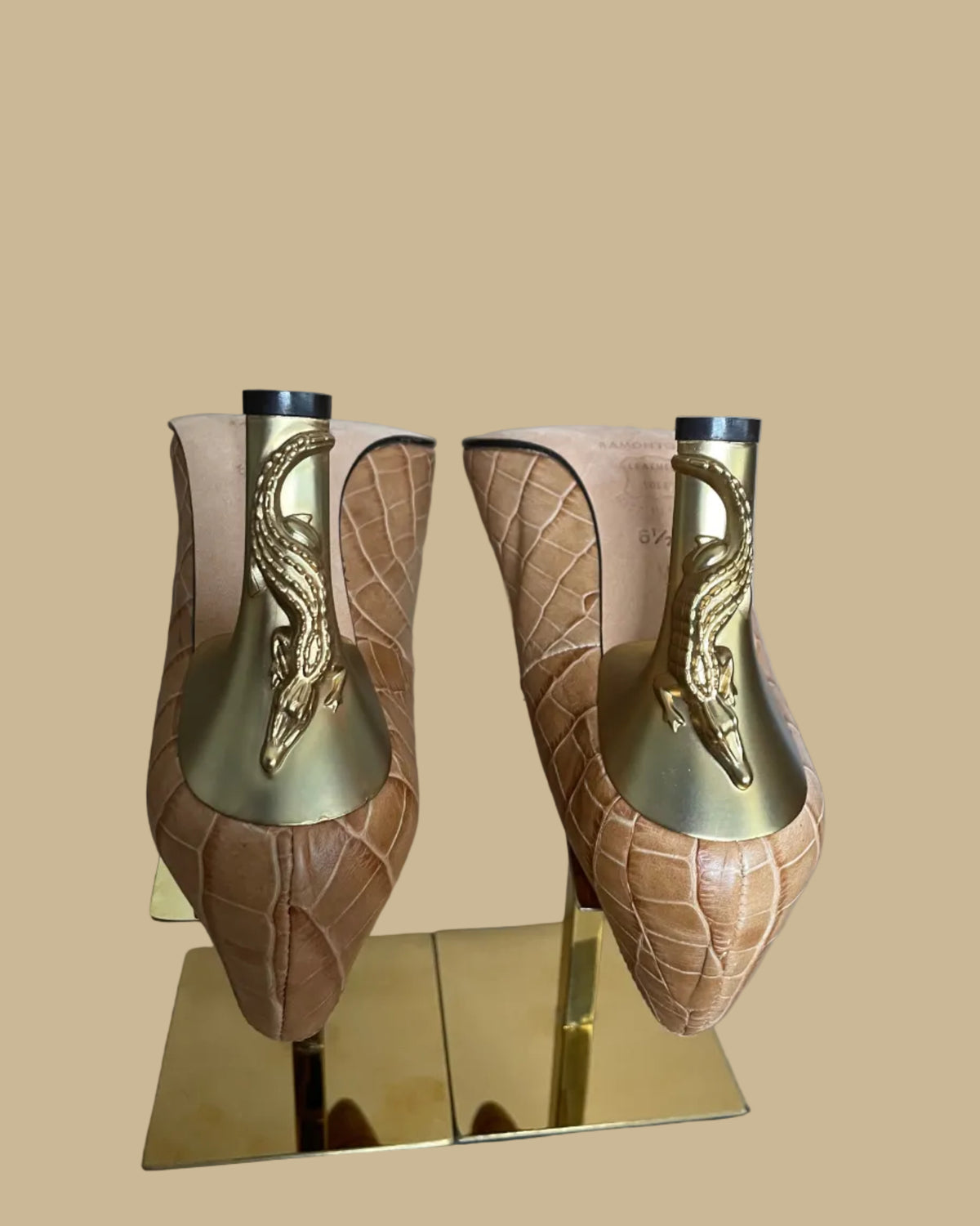 A pair of pointed-toe high heels in a tan crocodile-embossed leather, displayed on a gold-colored stand against a neutral beige background. The brand name Ramon Tenza is visible on the black insoles.
