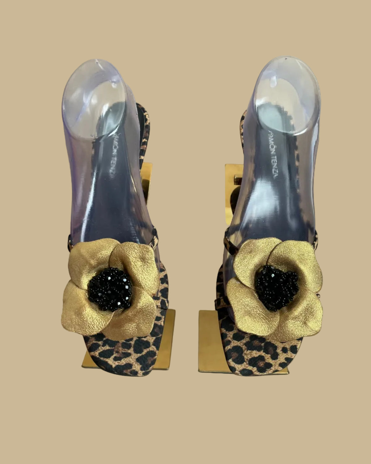 A pair of leopard print Ramón Tenza slingback sandals with a gold leather flower and black beaded center, displayed on gold stands against a kraft background