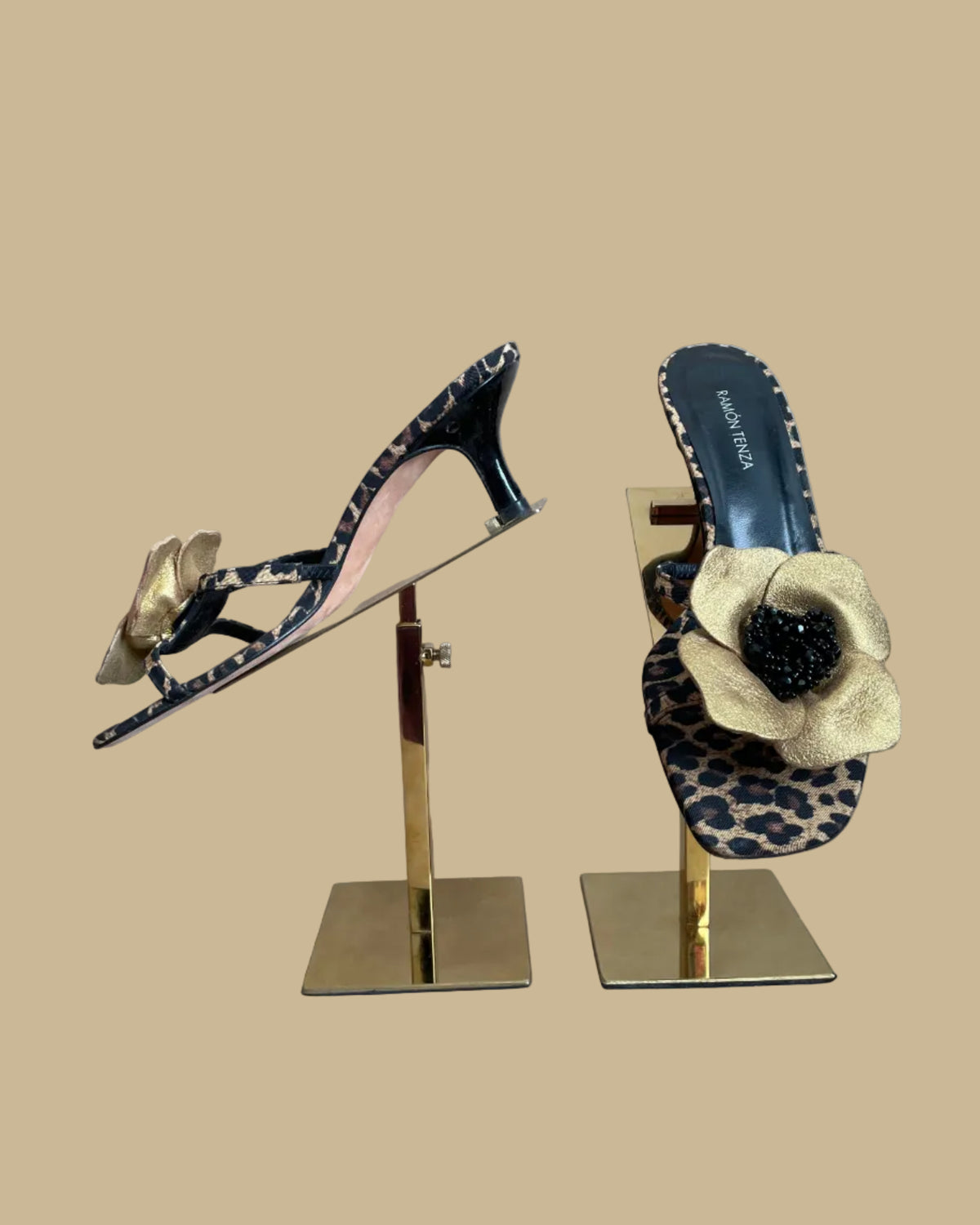 A pair of leopard print Ramón Tenza slingback sandals with a gold leather flower and black beaded center, displayed on gold stands against a kraft background