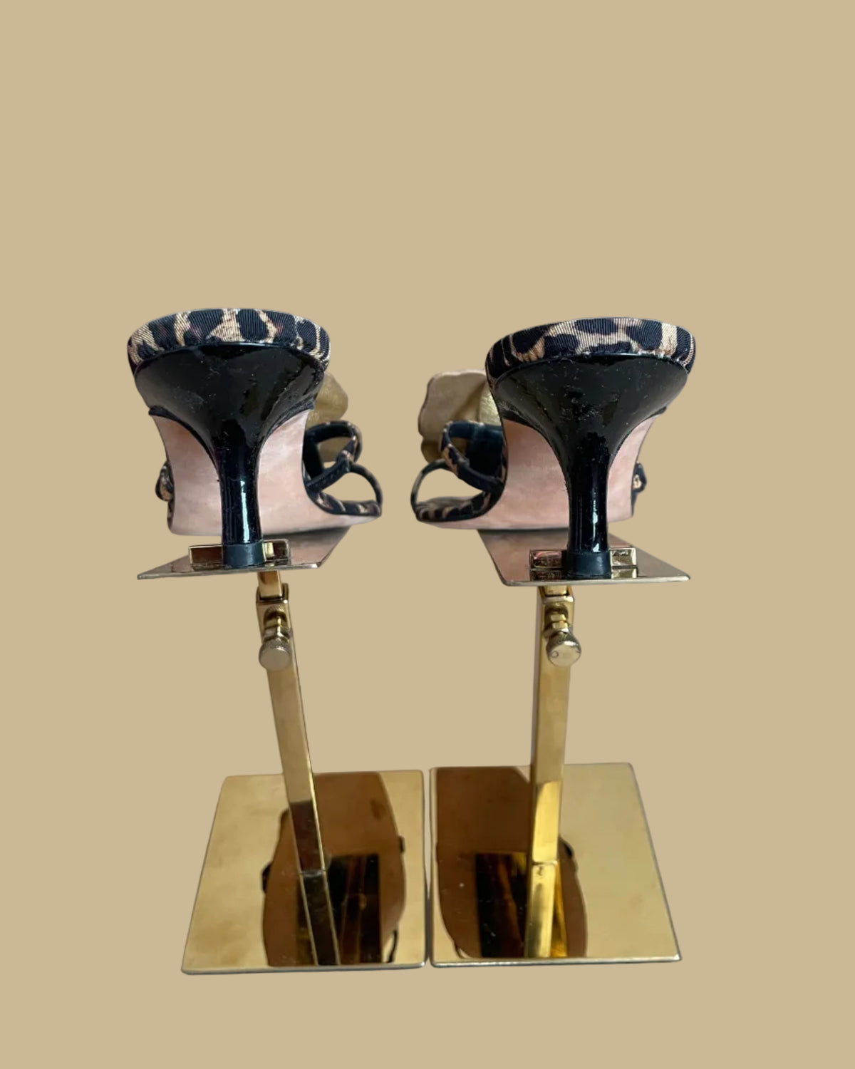 A pair of leopard print Ramón Tenza slingback sandals with a gold leather flower and black beaded center, displayed on gold stands against a kraft background