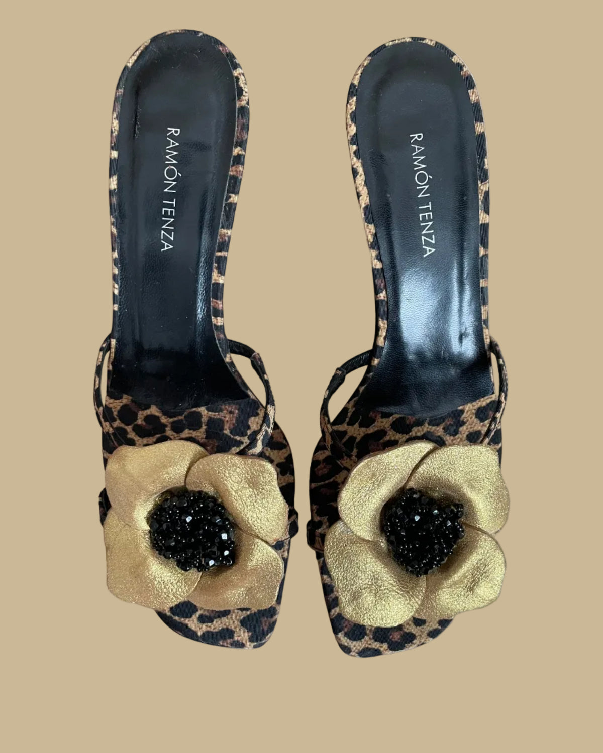 A pair of leopard print Ramón Tenza slingback sandals with a gold leather flower and black beaded center, displayed on gold stands against a kraft background