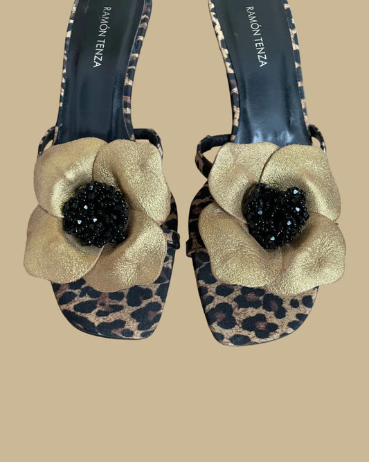 A pair of leopard print Ramón Tenza slingback sandals with a gold leather flower and black beaded center, displayed on gold stands against a kraft background
