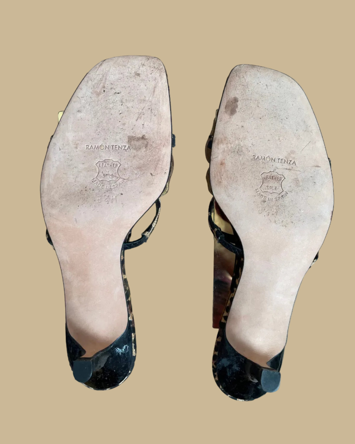 A pair of leopard print Ramón Tenza slingback sandals with a gold leather flower and black beaded center, displayed on gold stands against a kraft background