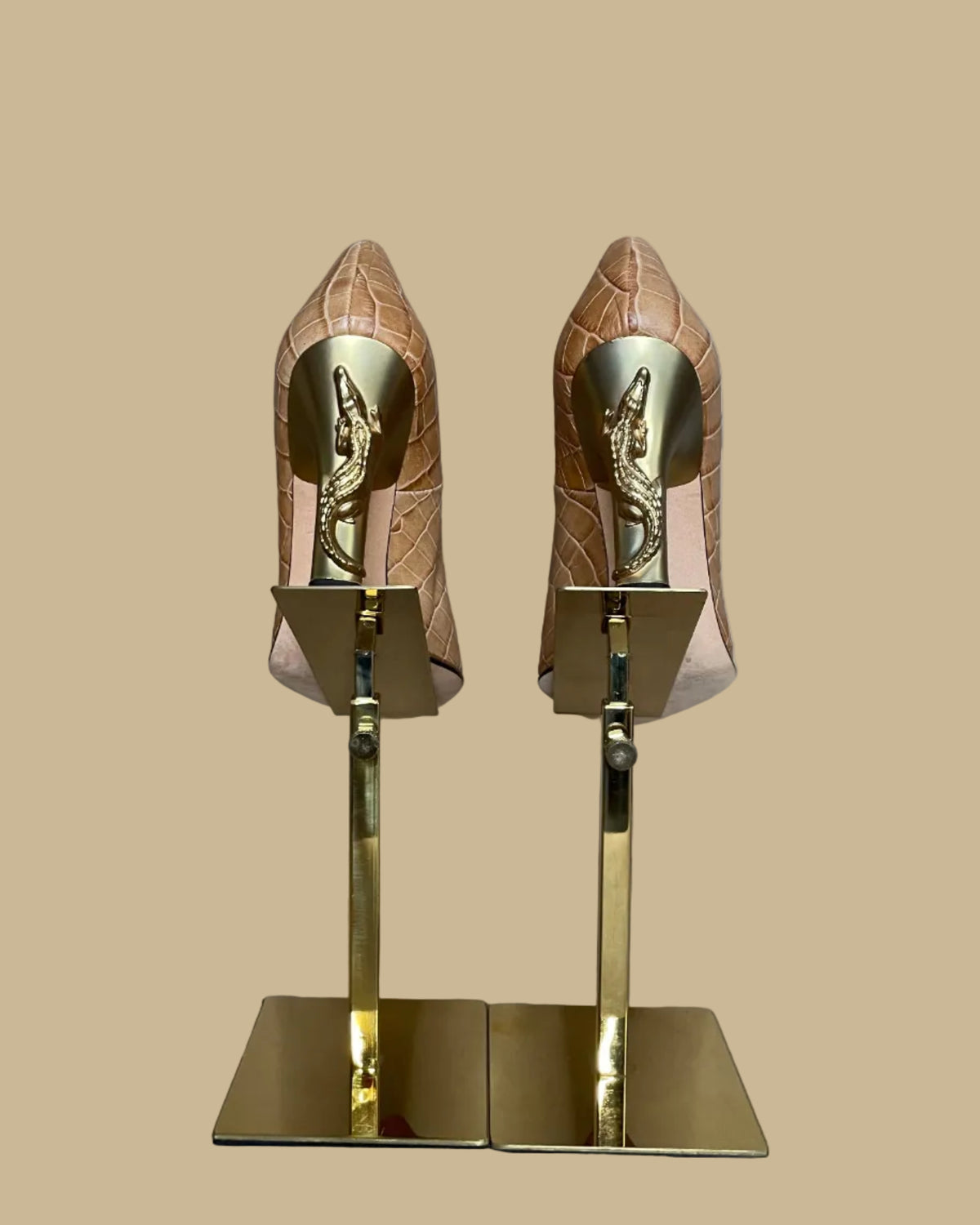 A pair of pointed-toe high heels in a tan crocodile-embossed leather, displayed on a gold-colored stand against a neutral beige background. The brand name Ramon Tenza is visible on the black insoles.