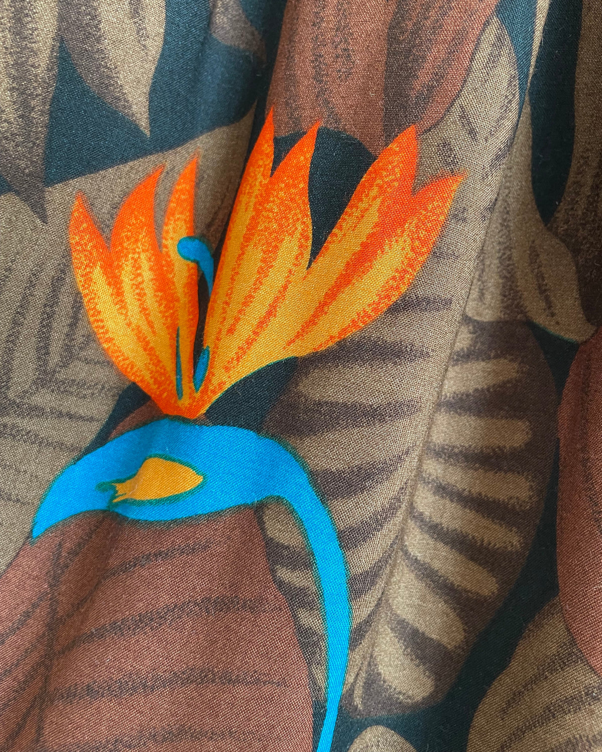 A vintage tropical bird-of-paradise print pleated skirt in brown, black, teal, and orange tones, shown against a kraft background
