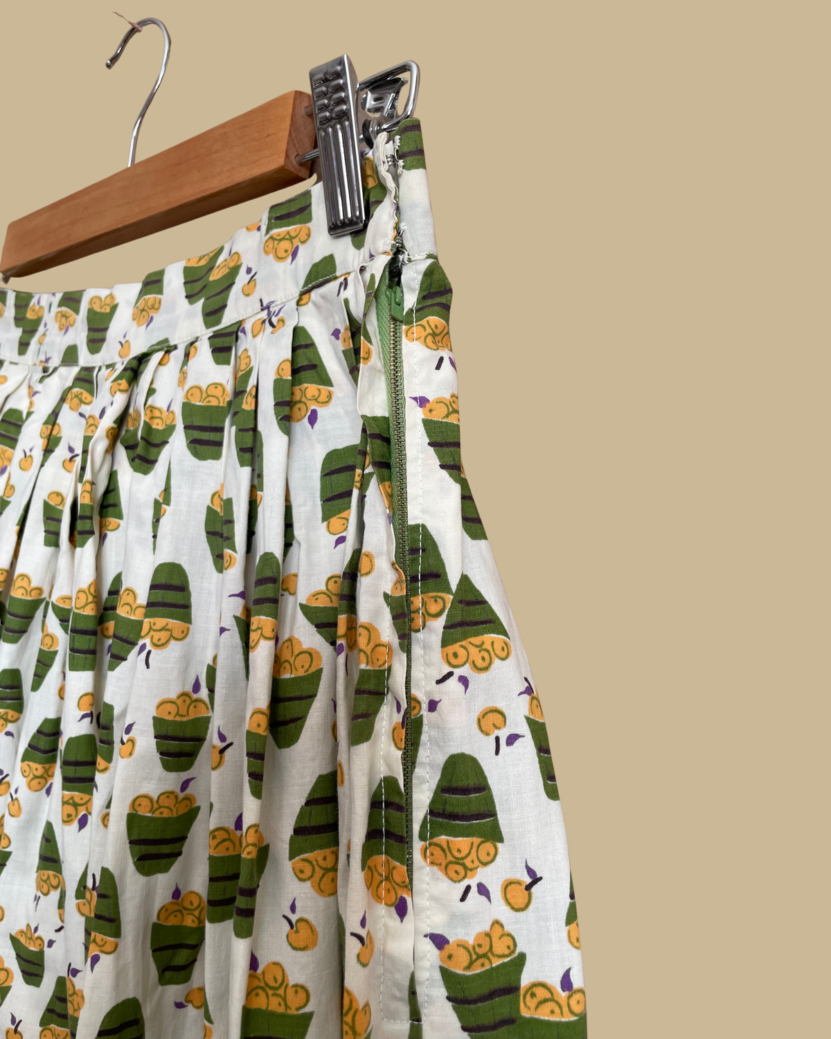 A vintage handmade pleated midi skirt with an apple basket novelty print in olive, mustard, and cream, shown against a kraft background