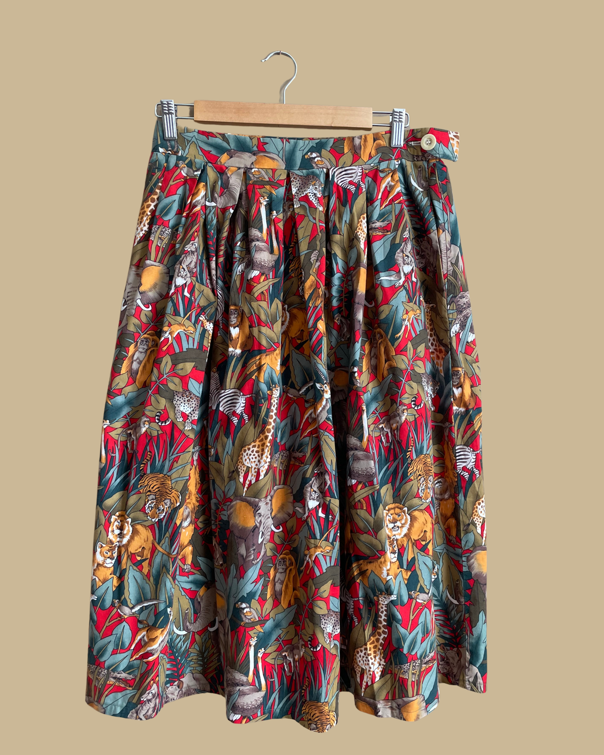 A vintage handmade pleated midi skirt with a jungle safari animal print featuring elephants, giraffes, zebras, and tigers in red, olive, and teal, shown against a kraft background