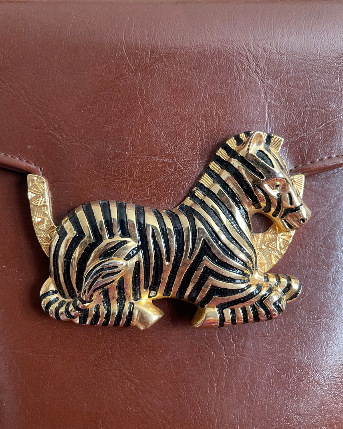 A vintage brown crossbody bag with a sculptural gold-tone and black enamel zebra clasp on the flap closure, shown against a kraft background
